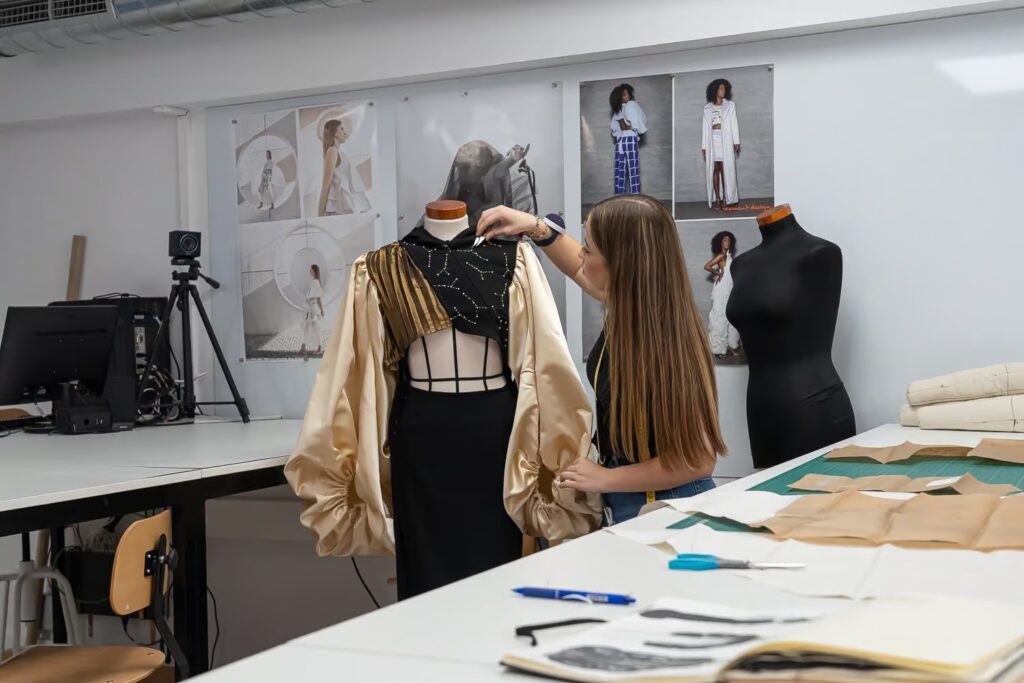 En la imagen se ve a una chica de pie, completamente concentrada mientras acomoda cuidadosamente un vestido sobre un maniquí. Está en medio de un estudio lleno de elementos relacionados con la moda: telas de distintos colores, tijeras, alfileres, bocetos colgados en las paredes y revistas abiertas en páginas de pasarela. Su mirada está fija en cada detalle del vestido, demostrando su amor por la moda y su dedicación a este arte.
La chica viste de forma moderna, reflejando su pasión por la moda no solo en lo que crea, sino también en cómo se expresa a través de su estilo personal. Cada movimiento que hace, desde alisar la tela hasta ajustar las costuras, revela su profundo conocimiento de la moda y su deseo de innovar dentro del mundo de la moda. En este instante, ella no solo está confeccionando una prenda: está dando vida a una idea, a una visión única de lo que para ella significa la moda. Es un momento donde la creatividad y la moda se encuentran cara a cara frente al maniquí.